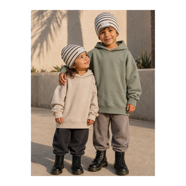 Two children standing outdoors wearing matching knit beanie hats and hoodies. The child on the right wears a muted sage green hoodie and gray joggers with black boots, while the child on the left wears a light beige hoodie and dark gray pants.