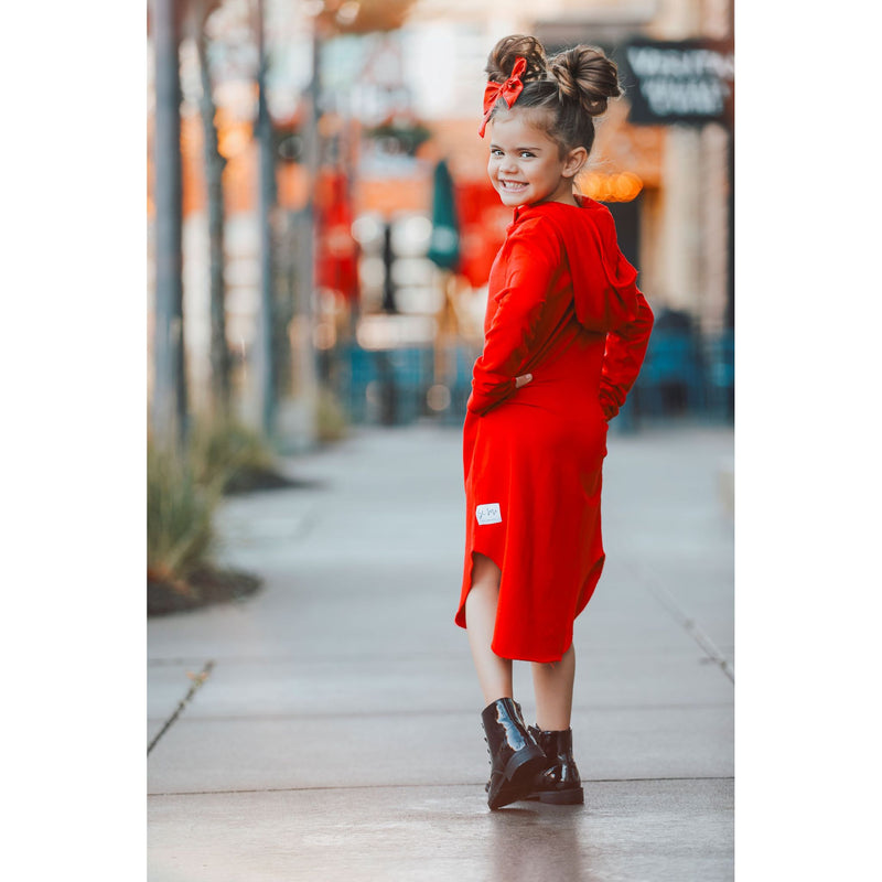 Young girl wearing a bright red hooded dress, smiling over her shoulder while standing on a city sidewalk, showing the dress’s back and curved hemline