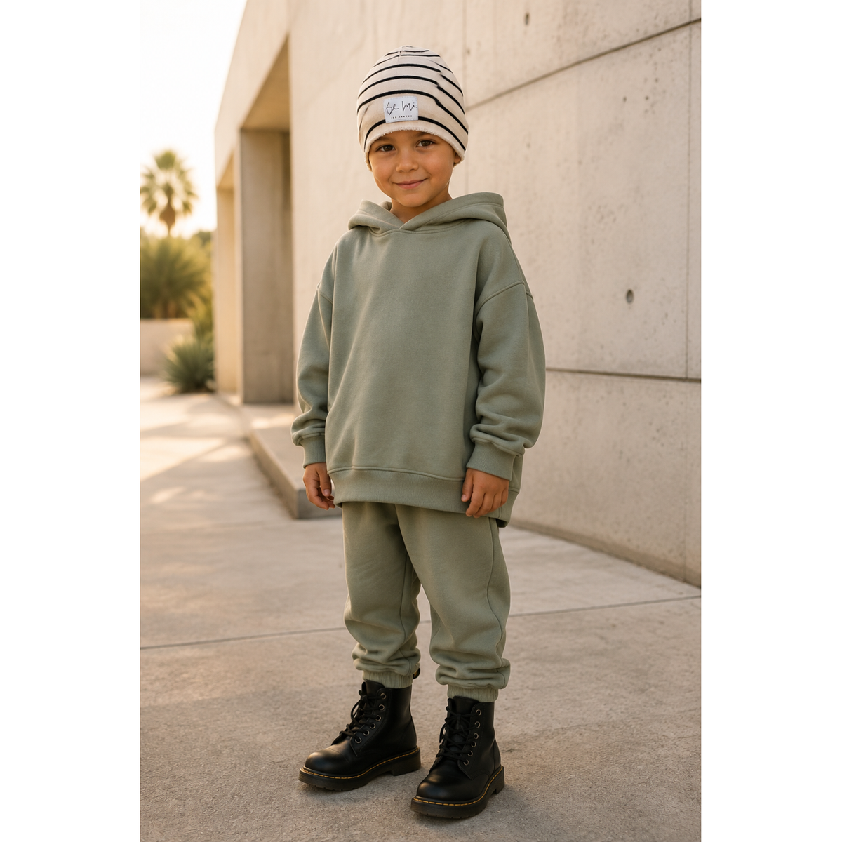 A young child wearing a matching sage green hoodie and jogger sweatpants set, a black and white striped beanie, and black boots, standing outdoors against a concrete wall.