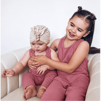 Two young girls wearing matching dusty rose one-piece outfits sit together on a cream-colored armchair against a plain white wall. The older girl has her hair in two buns and is smiling, while the younger girl wears a matching beige fabric headwrap.