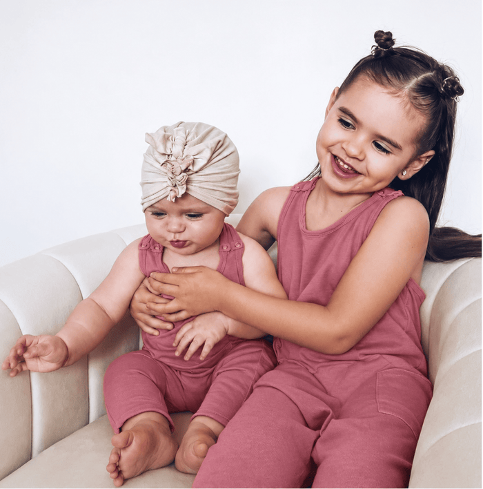 Two young girls wearing matching dusty rose one-piece outfits sit together on a cream-colored armchair against a plain white wall. The older girl has her hair in two buns and is smiling, while the younger girl wears a matching beige fabric headwrap.