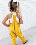 A toddler girl seen from behind wearing a mustard yellow sleeveless jumpsuit over a white tank top, with black over-ear headphones on. She is standing on a concrete surface against a white brick wall.