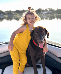 A young girl in a bright yellow sleeveless jumpsuit stands on a boat next to a brown dog wearing a red collar, with water and trees in the background.
