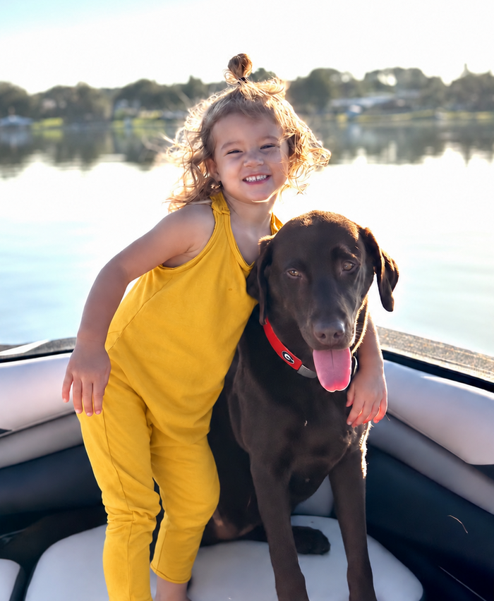 A young girl in a bright yellow sleeveless jumpsuit stands on a boat next to a brown dog wearing a red collar, with water and trees in the background.