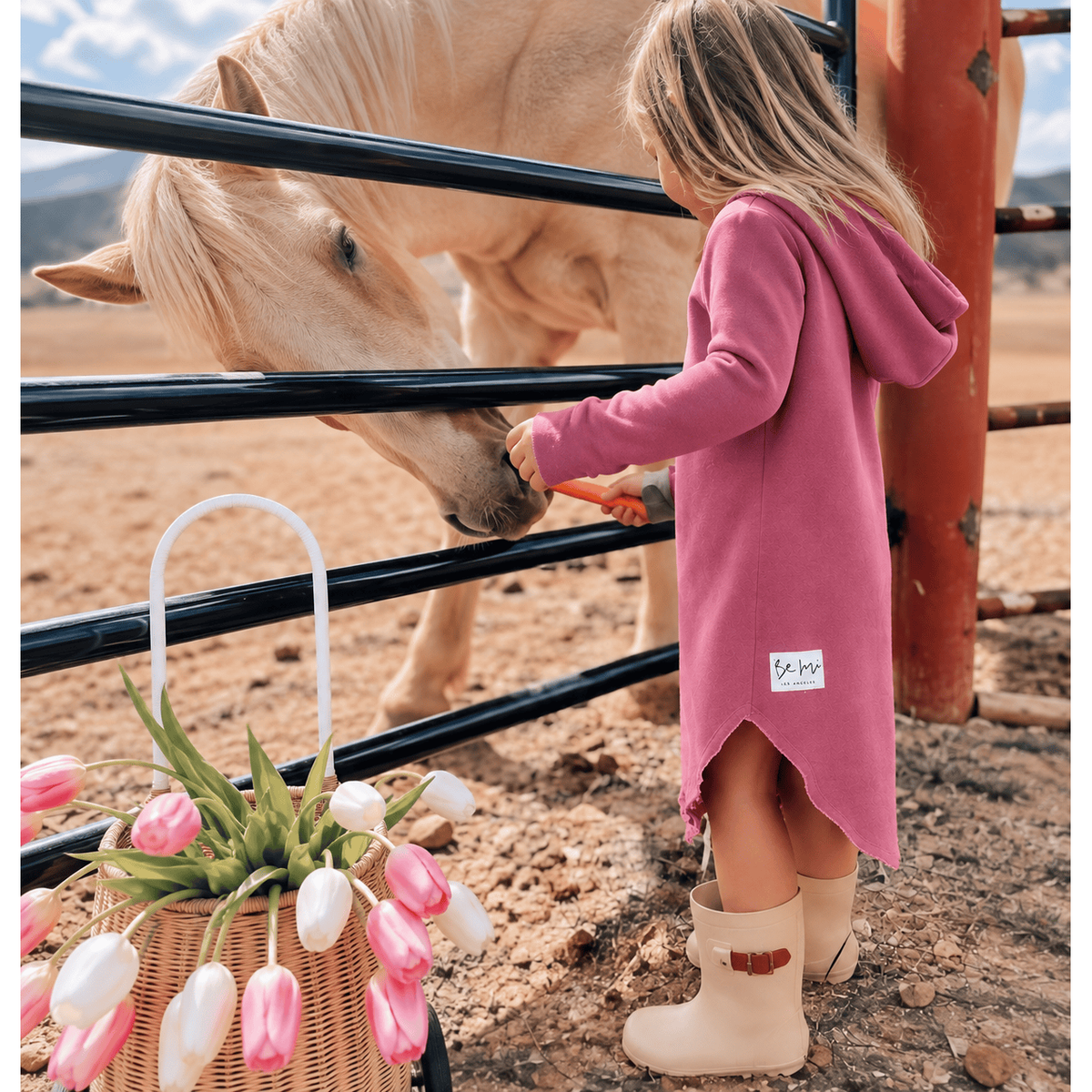 A young girl in a dusty pink hooded dress and beige rain boots feeds a light-colored horse through a black metal fence in an outdoor ranch setting. A basket of pink and white tulips sits in the foreground.