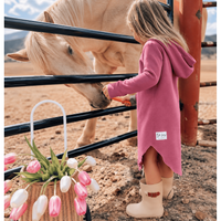 A young girl in a dusty pink hooded dress and beige rain boots feeds a light-colored horse through a black metal fence in an outdoor ranch setting. A basket of pink and white tulips sits in the foreground.