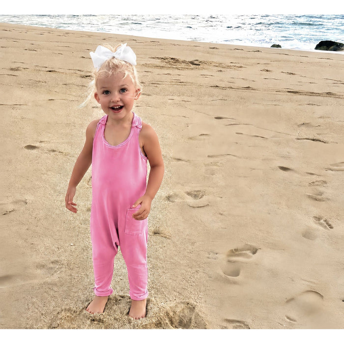 A young toddler girl with blonde hair in a white bow stands on a sandy beach wearing a pink sleeveless overall jumpsuit.