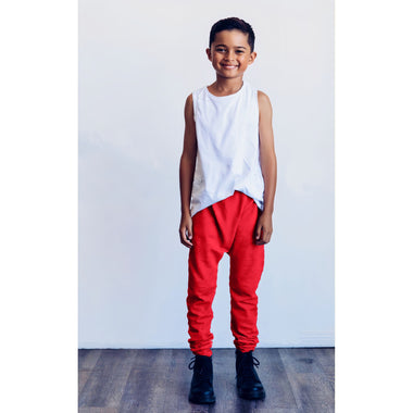 Child wearing a white sleeveless shirt and red pants standing against a white wall.