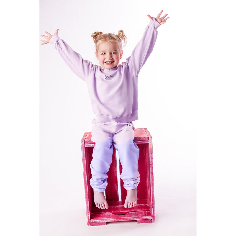 Child in a purple outfit sitting on a pink box with arms raised against a white background