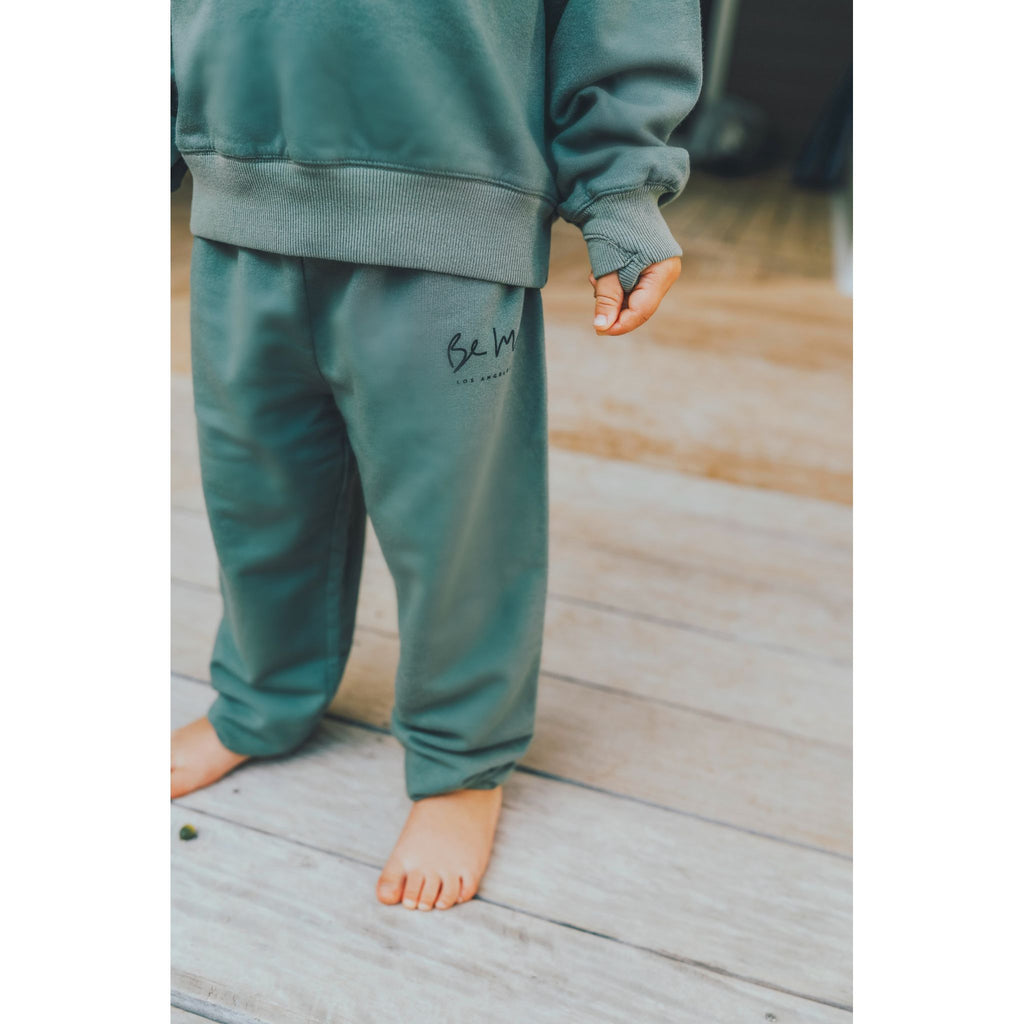 Child wearing green organic sweatsuit an standing against a light gray wall.