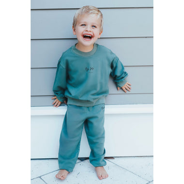 Child wearing green organic sweatsuit an standing against a light gray wall.